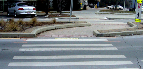 Photo shows a refuge island in the middle of the street, with a cut-through.  The cut-through has detectable warnings at each end, and starts heading across the street, turns to the right and then turns back to head straight across the street again.  The cut-through is perpendicular to the street for about 2 feet on each side of the island.